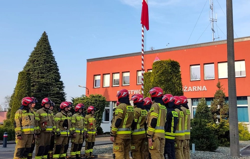 Strażacy w dwóch szeregach przed strażnicą.