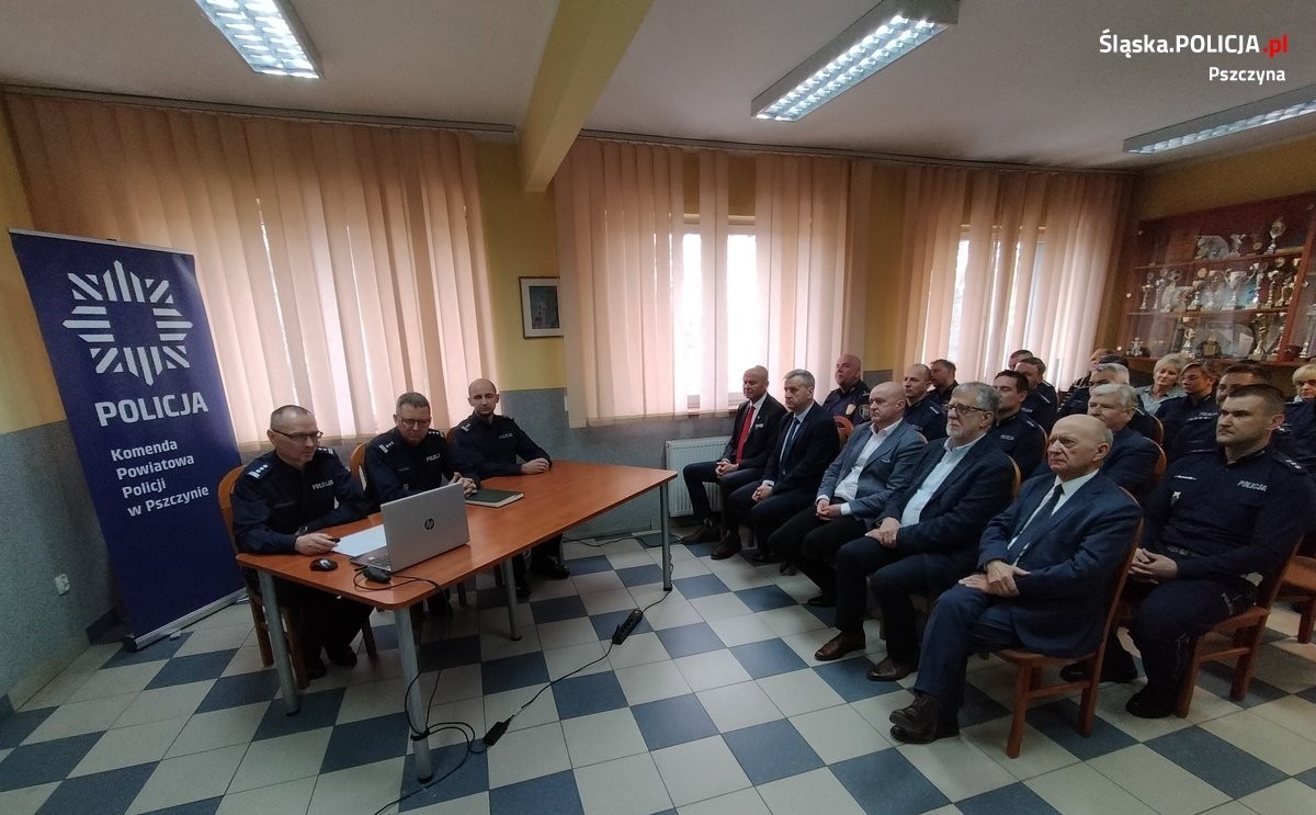 Przy stole siedzą policjanci prowadzący naradę, na przeciwko na krzesłach zaproszeni goście, w tym samorządowcy.