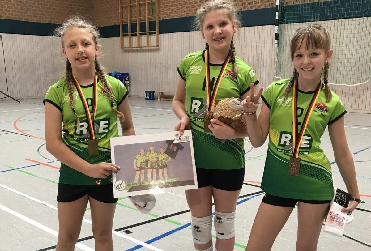 Trzy młode siatkarki w sportowych strojach stoją w hali sportowej
