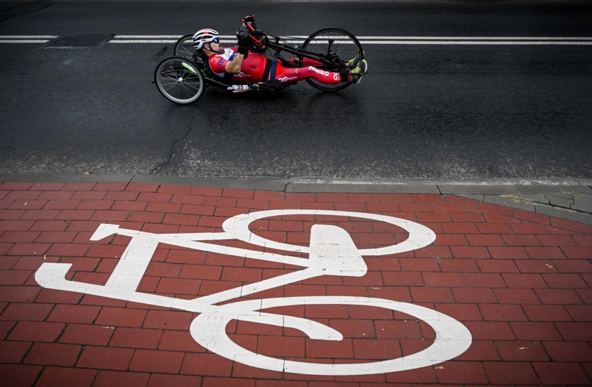 Niepełnosprawny sportowiec w czerwonym kombinezonie porusza się na rowerze typu hanbike po drodze. Na pierwszym planie na chodniku wymalowany sumbol wózka inwalidzkiego