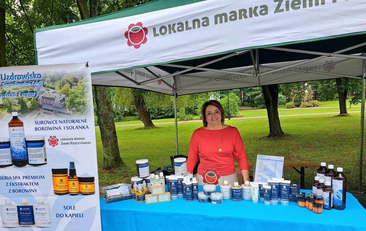 Kobieta stojąca pod namiotem lokalnej marki ziemi pszczyńskiej. Na stoisku produkty z uzdrowiska.