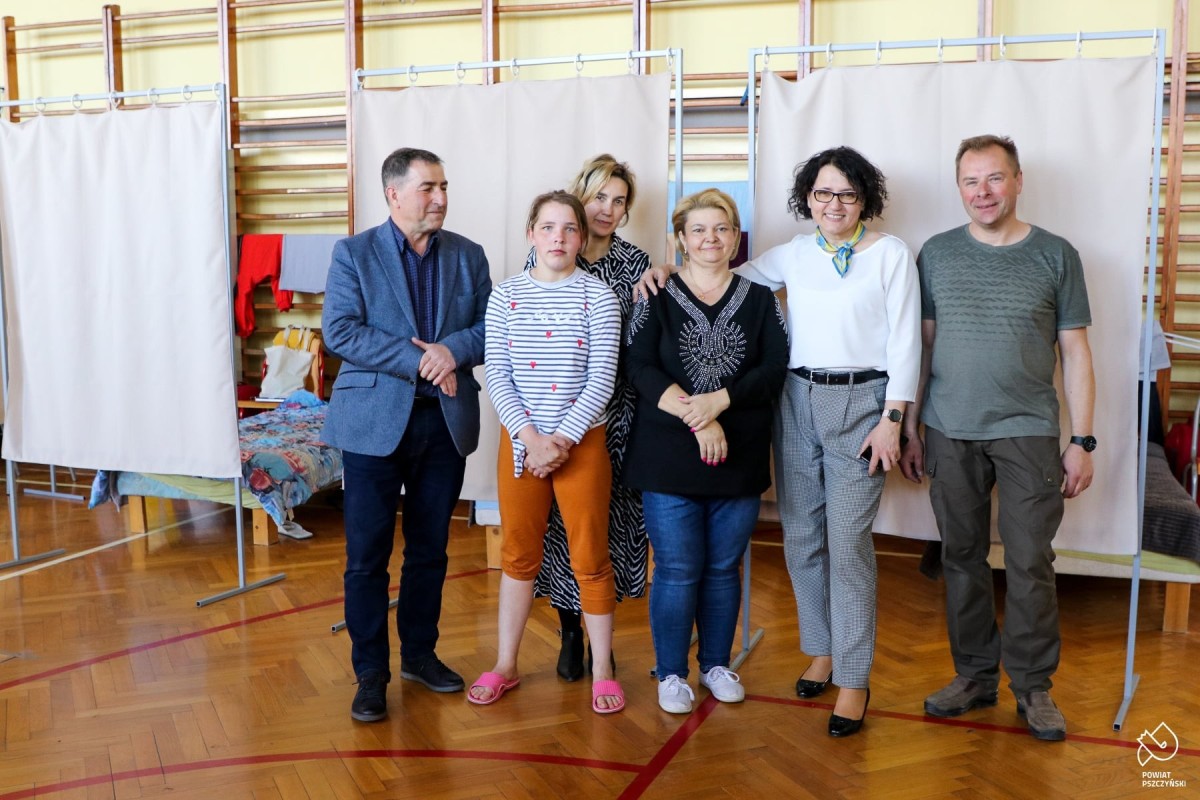 Grupa ludzi stoi w dużej sali, w której zorganizowano łóżka dla uchodźców