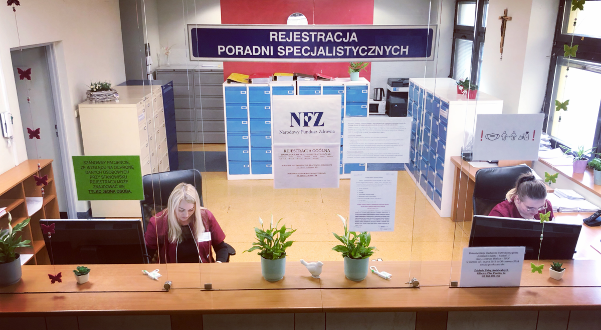 Rejestracja do poradni specjalistyczncyh w pszczyńskim szpitalu. Dwie rejestratorki siedzą za ladą w przeszklonym pomieszczeniu.