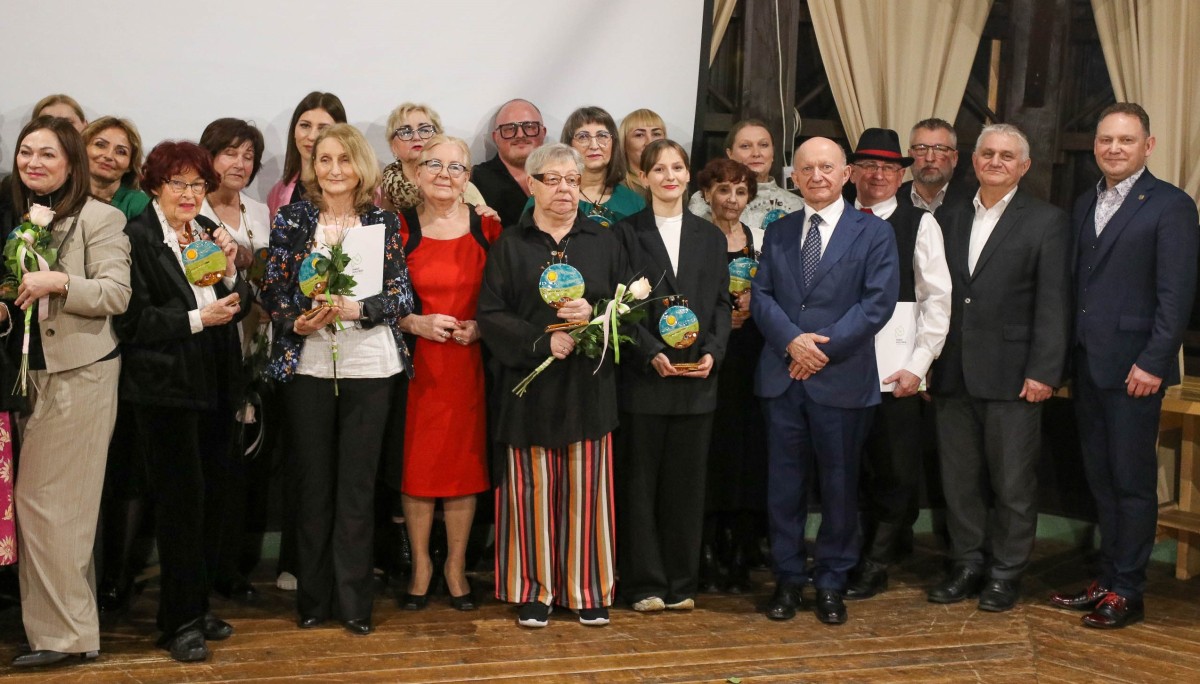 Duża grupa osób stoi obok siedzi, część trzyma statuetki, część dyplomy, a także białe róże.