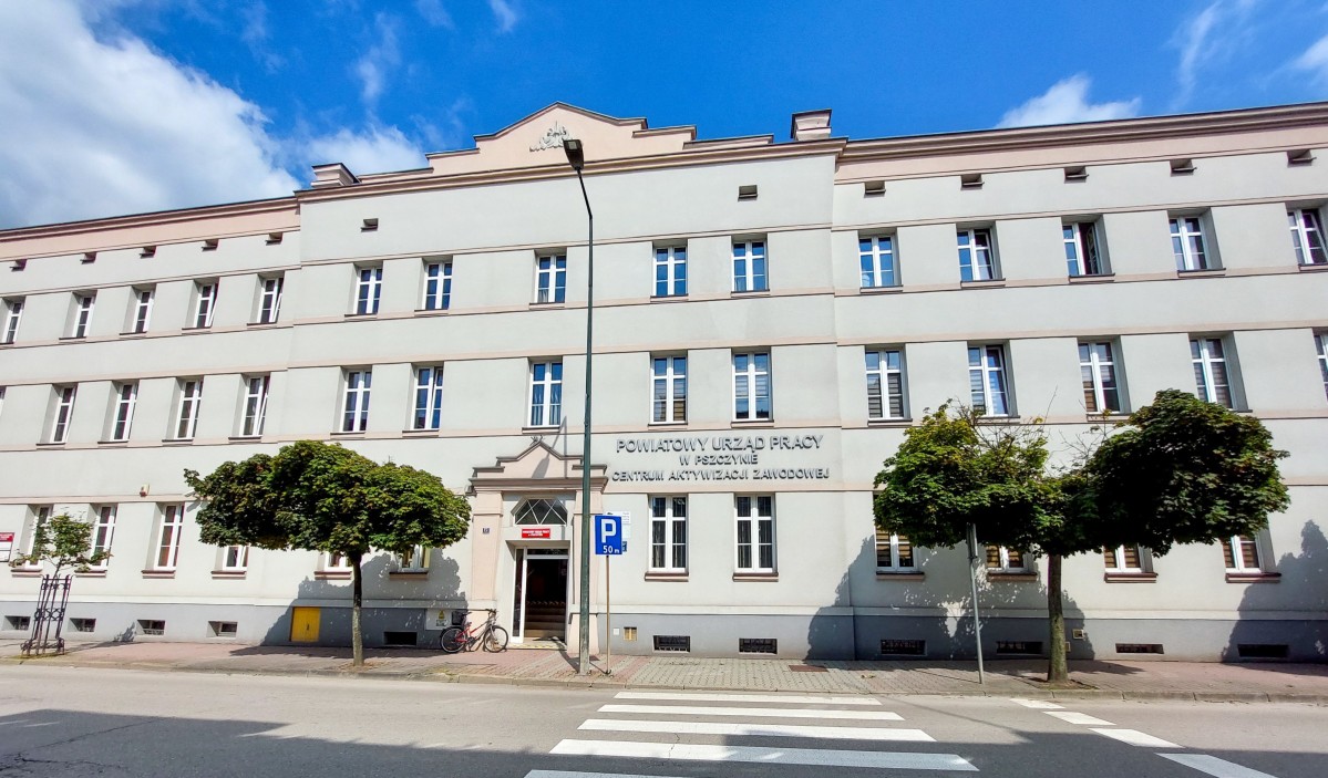 Budynek z licznymi oknami, na którym widnieje napis Powiatowy Urząd Pracy w Pszczynie. Do budynku o jasnej elewacji prowadzi przejście dla pieszych.