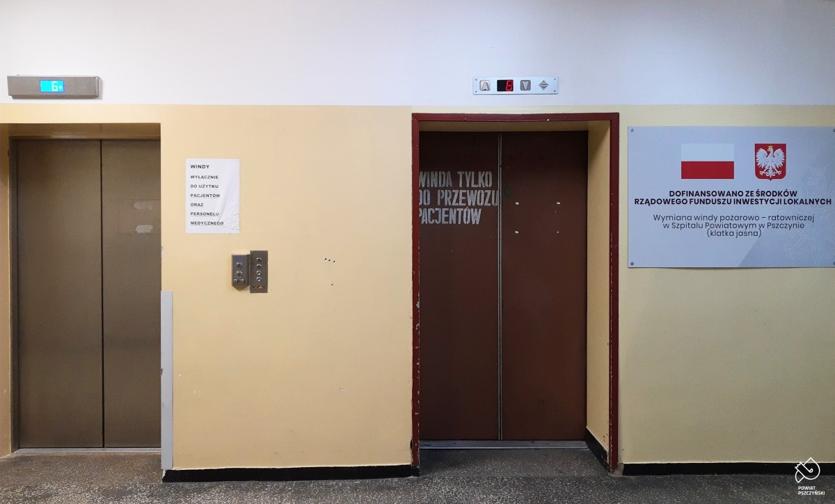 Szpitalny korytarz z dwoma windami. Jedna z nich jest nowa z nierdzewnymi drzwiami, a druga jest stara z brązowymi drzwiami z napisem "Winda tylko do przewozu pacjentów".