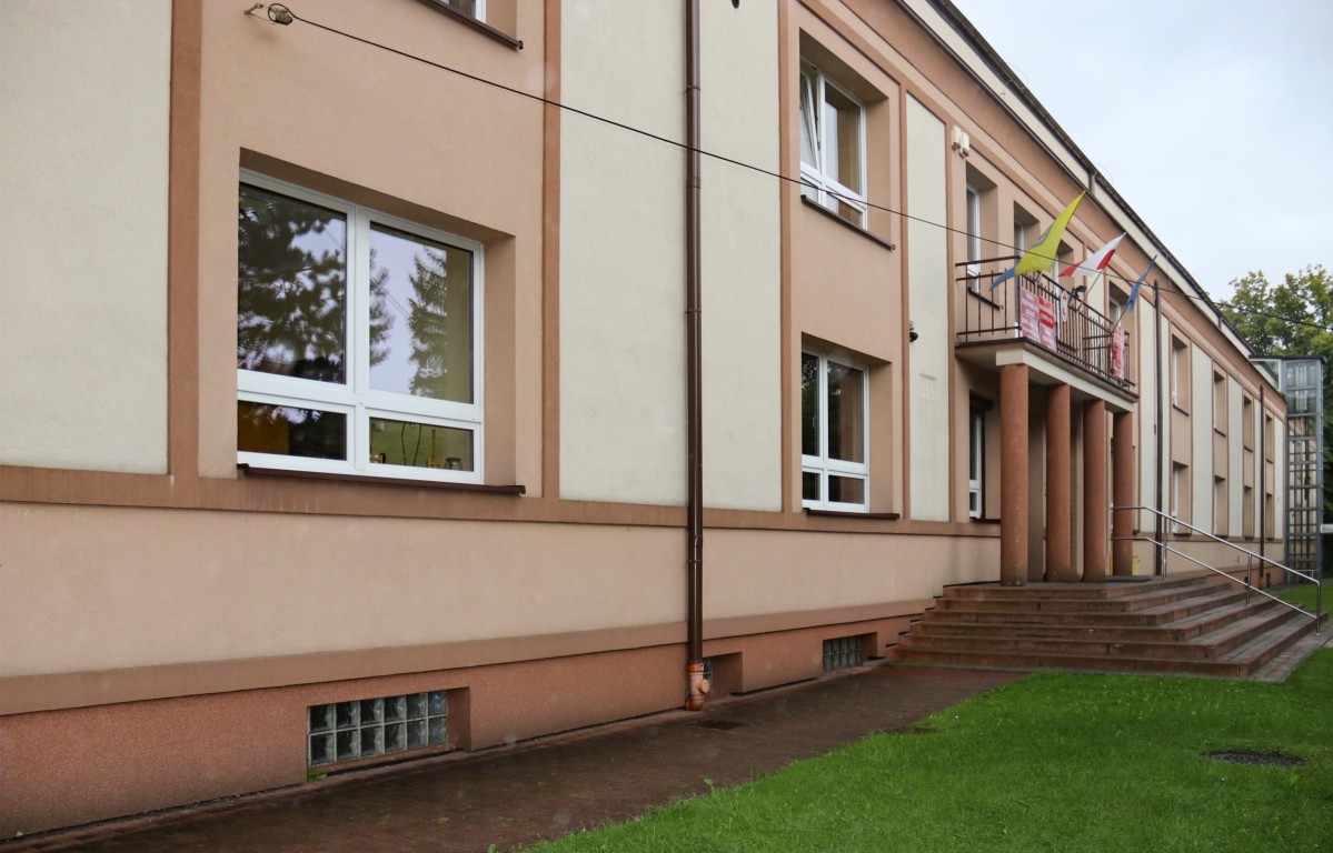 Jednopiętrowy brązowo-kremowy budynek z wywieszonymi nad wyjściem trzema flagami na balkonie.