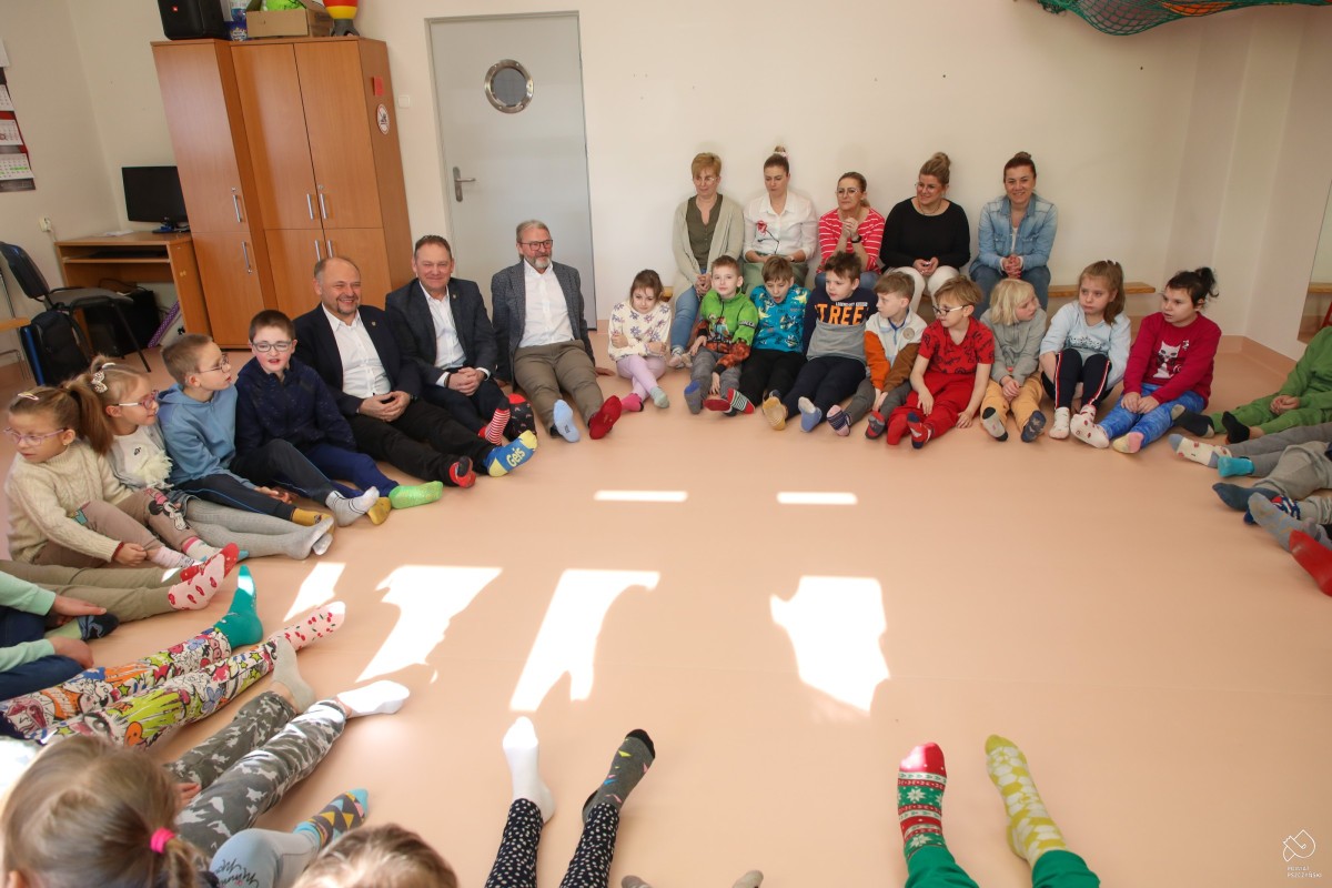 Starosta, wicestarosta i dyrektor szkoły siedzą z dziećmi w okręgu na ziemi. Na stopach wszyscy mają kolorowe skarpetki.