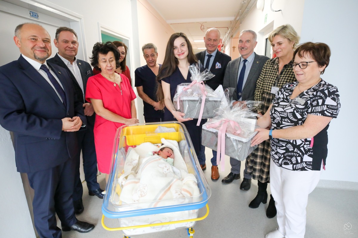 pracownicy szpitala i goście ze starostwa stoją na korytarzu razem z mamą pierwszej dziewczynki urodzonej w 2025 r. w pszczyńskim szpitalu. Przed nimi stoi przeźroczyste łóżeczko, w którym lezy dziewczynka. Ma bujne czarne włosy