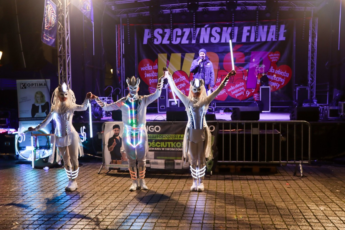 Trzy osoby w ledowych strojach, kłaniają się, za nimi scena a na niej prowadzący.