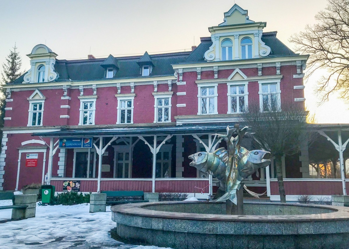 Budynek z jasno zieloną elewacją, na nim napis szpital. Przed nim rosną wysokie drzewa