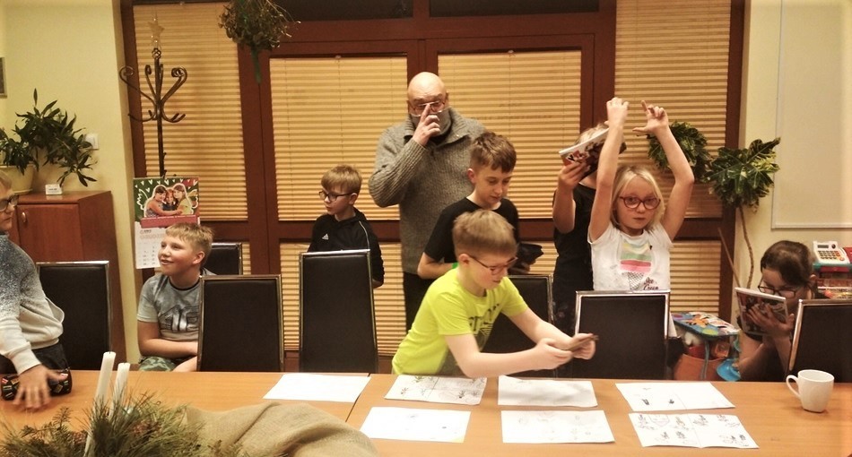Grupa dzieci stojących przy stole z mężczyzną w okularach prowadzącym warsztaty.