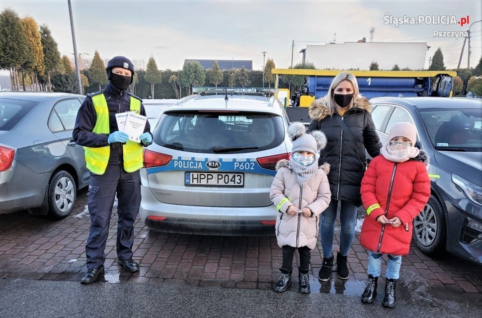 Policjant i kobita z dwójką dzieci stojący przy radiowozie. Na twarzach mają założone maseczki, a na kurtkach odblaski.