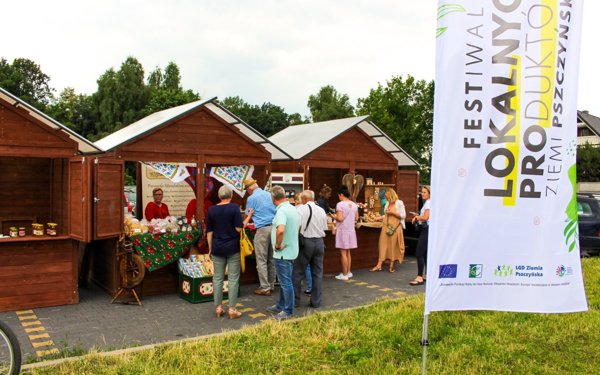 Drewniane stoiska z lokalnymi produktami, przed którymi stoją zainteresowani mieszkańcy i turyści.