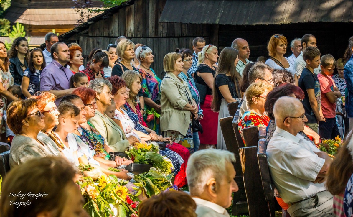 Ludzie siedzący na krzesłach podczas mszy polowej w skansenie, w dłoniach trzymają bukiety z ziół i kwiatów.