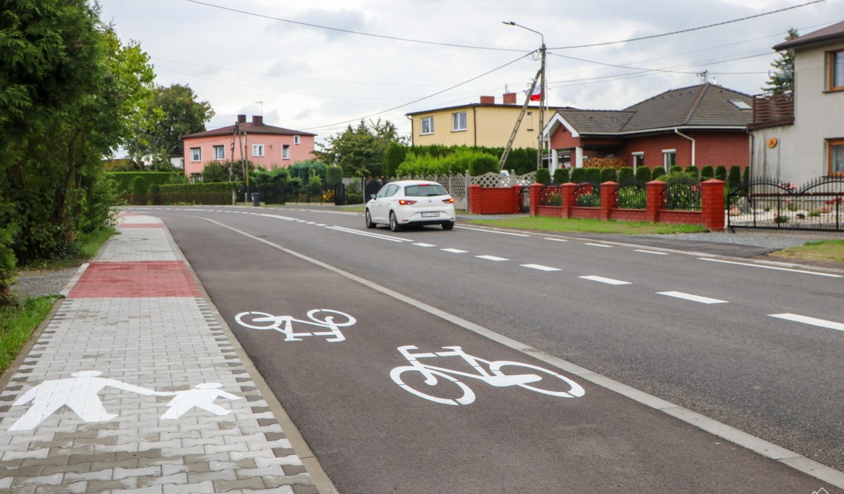 Wyremontowana nawierzchnia drogi, wraz z chodnikiem i ścieżką rowerową. Drogą jedzie srebrny samochód