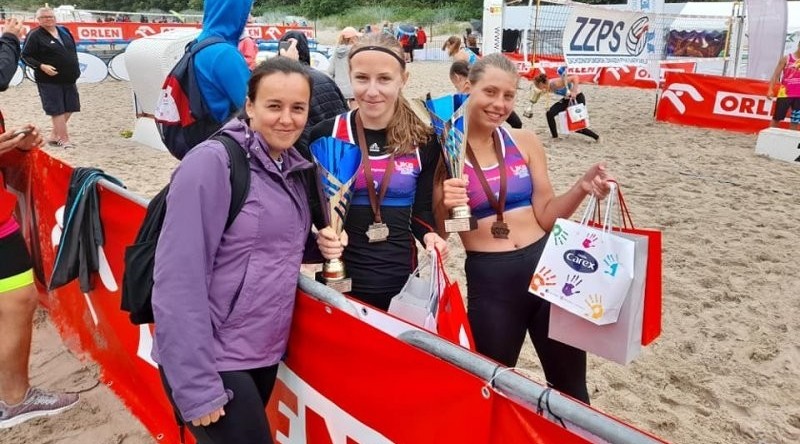 Na boisku plażowym stoją dwie młode siatkarki wraz z trenerką.