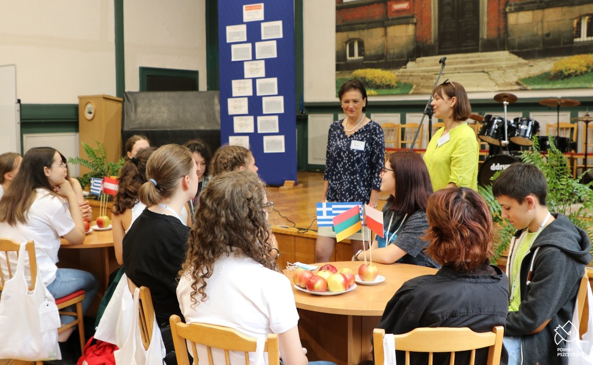 Uczniowie wraz z nauczycielami siedzący przy okrągłych stołach. Przed sceną stoi dyrektor placówki oraz koordynatorka projektu Erasmus+.