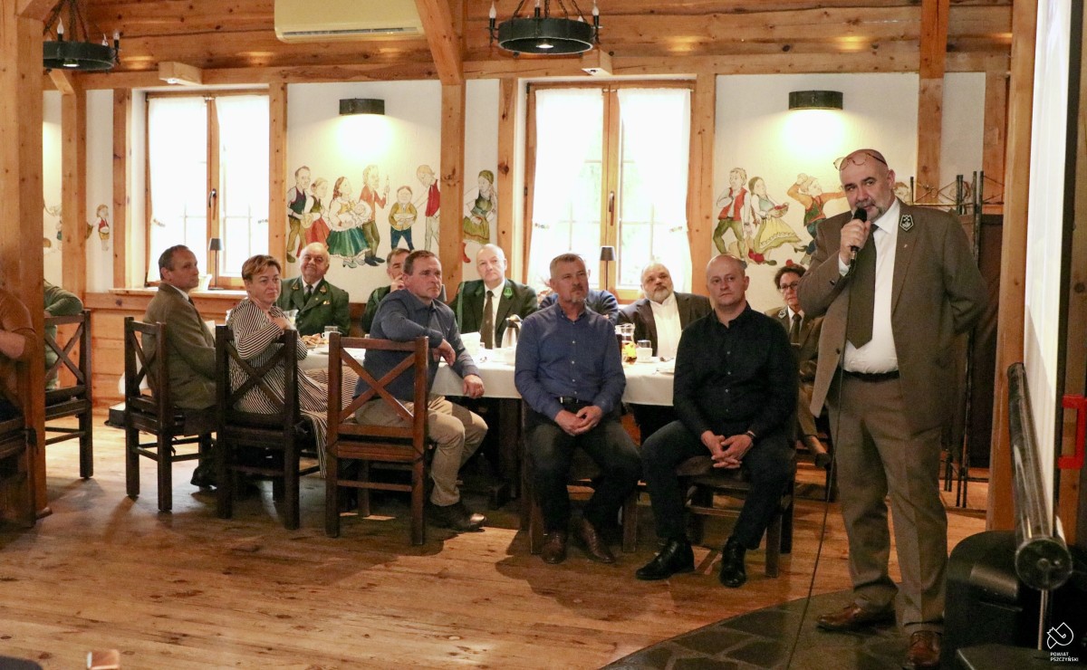 P.o. nadleśniczego przemawia, trzyma mikrofon, w tle siedzą na krzesłach przy stole w restauracji uczestnicy.