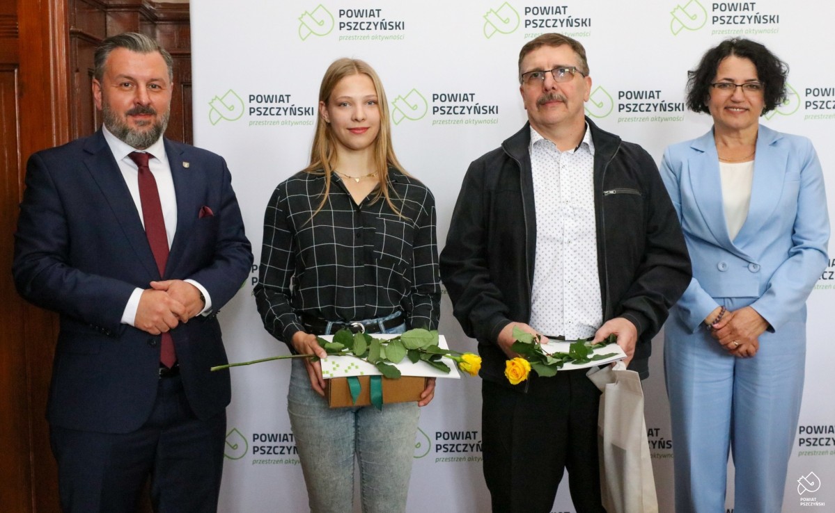 Zawodniczka z trenerem, starostą i przewodniczący Rady we wspólnym zdjęcie na ściance z logo powiatu.