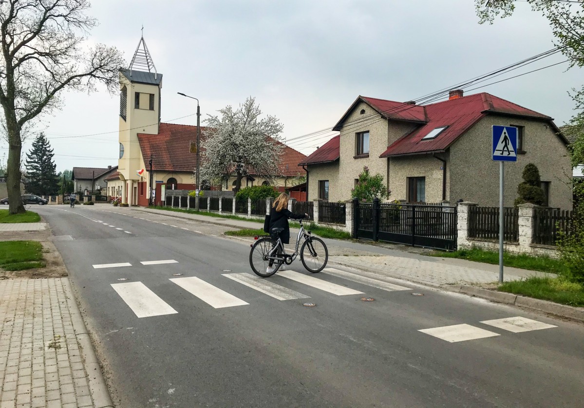 Przejście dla pieszych przez drogę, po którym przechodzi kobieta prowadząca rower. W oddali budynek kościoła.