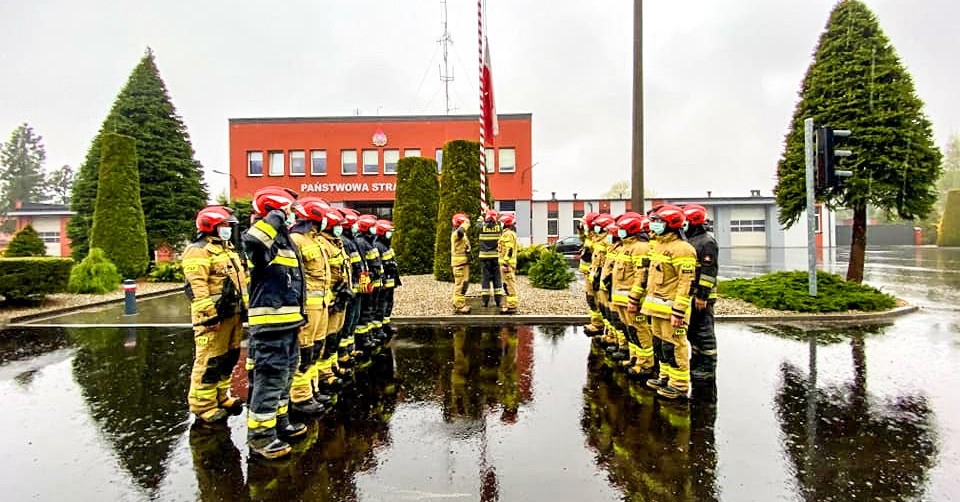 Strażacy stojący w dwóch rzędach na przeciwko siebie. W tle budynek komendy straży pożarnej w Pszczynie. Pada deszcz.