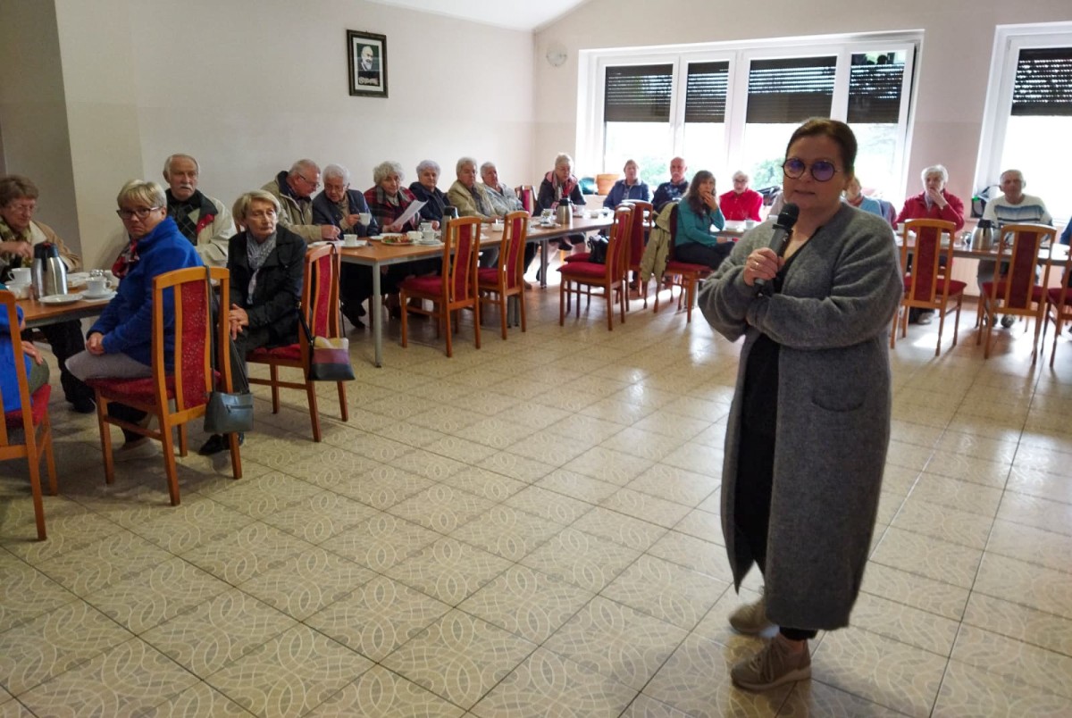 Rzecznik konsumentów stoi pośrodku sali, gdzie przy stołach siedzą seniorzy.