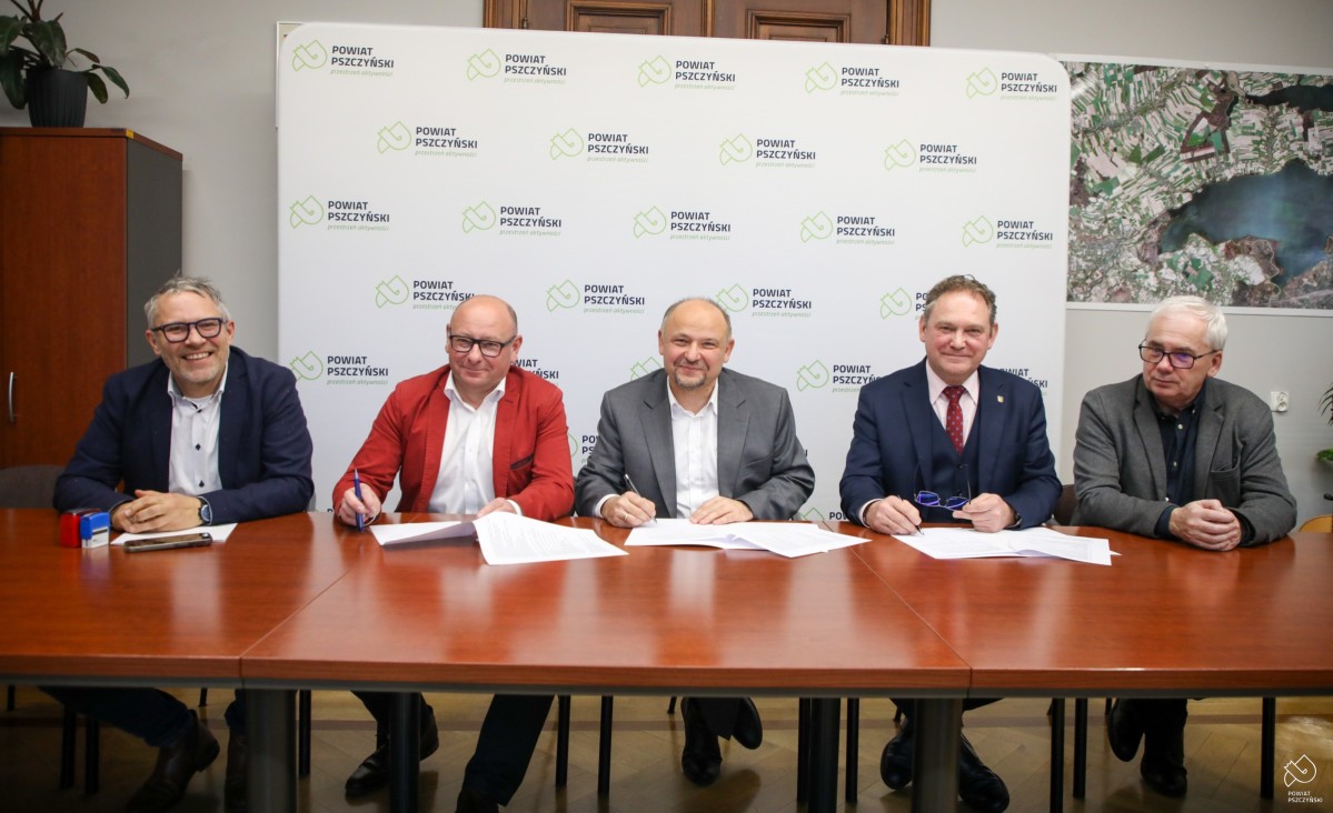 Kilka mężczyzn siedzi za stołem w gabinecie starosty, za nimi ścianka z logiem Powiatu.