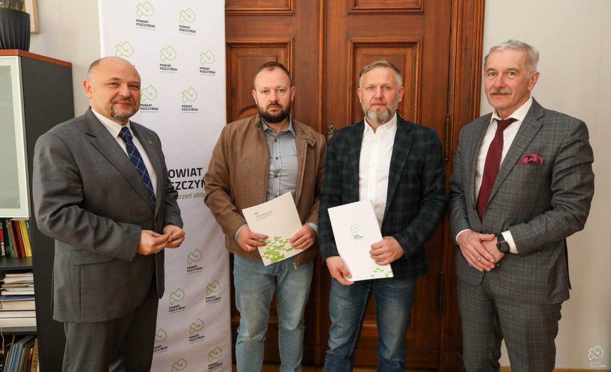 Cztery osoby na tle drzwi i ścianki powiatu, dwie osoby trzymają teczki z umową.