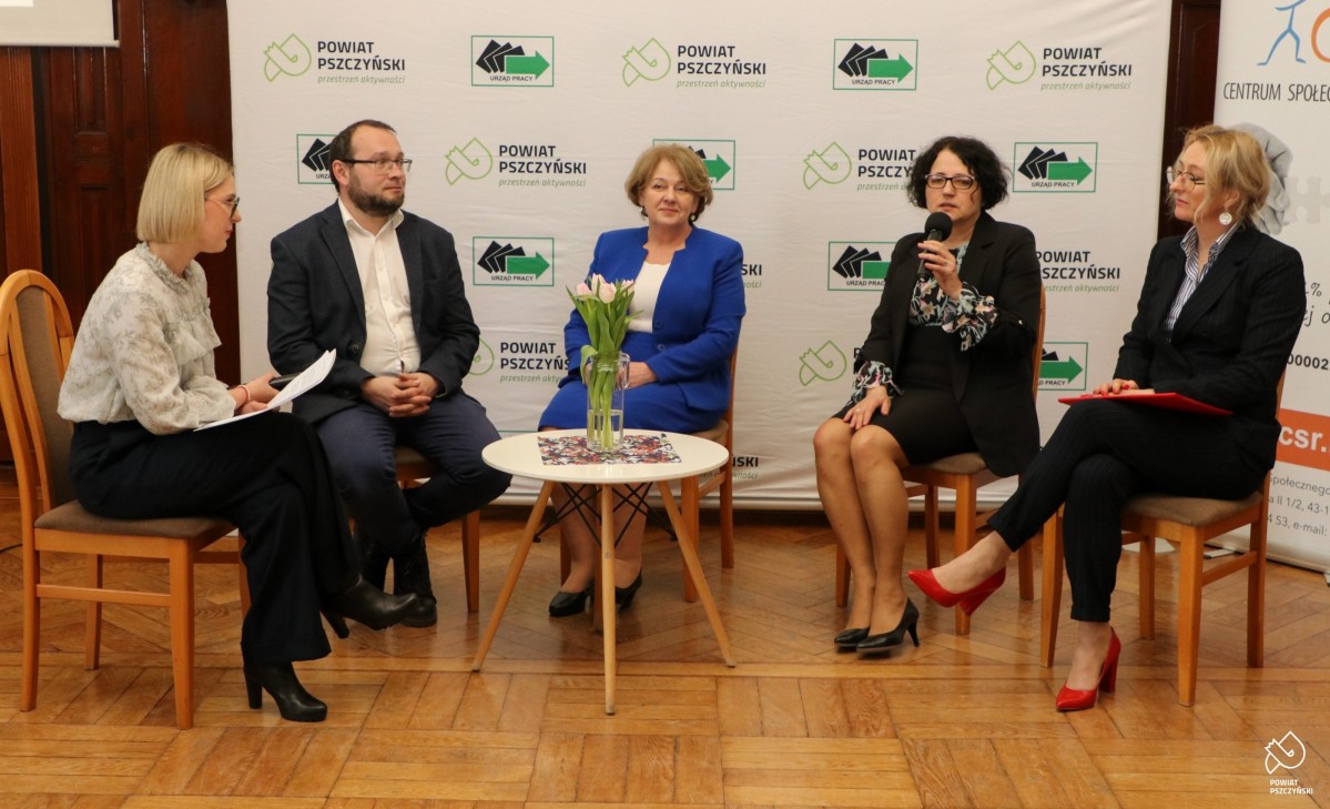 Osoby biorące udział w panelu dyskusyjnym siedzą na krzesłach w sali sesyjnej, rozmawiają.