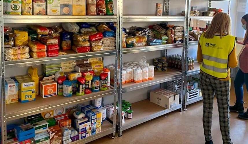 pomieszczenie z regałami, na których ustawiono produkty żywnościowe. Przed regałem stoi wolontariuszka w żółtej kamizelce.