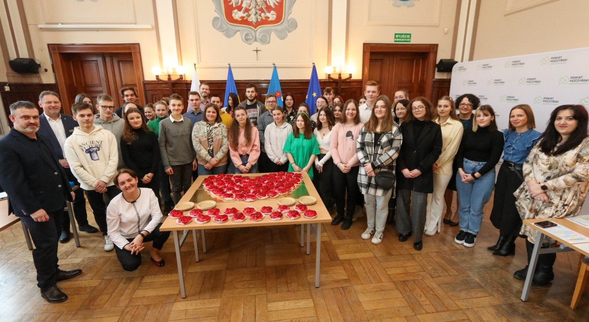 Grupa wolontariuszy stoi w sali sesyjnej starosta. przed nimi na stole w kształcie serca ułożono babeczki