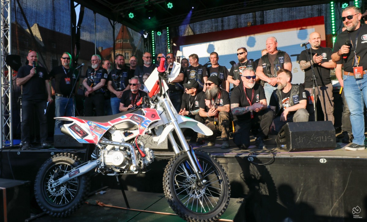 Organizatorzy motoserca w czarnych koszulkach z logo stoją na scenie, przed nimi stoi cross