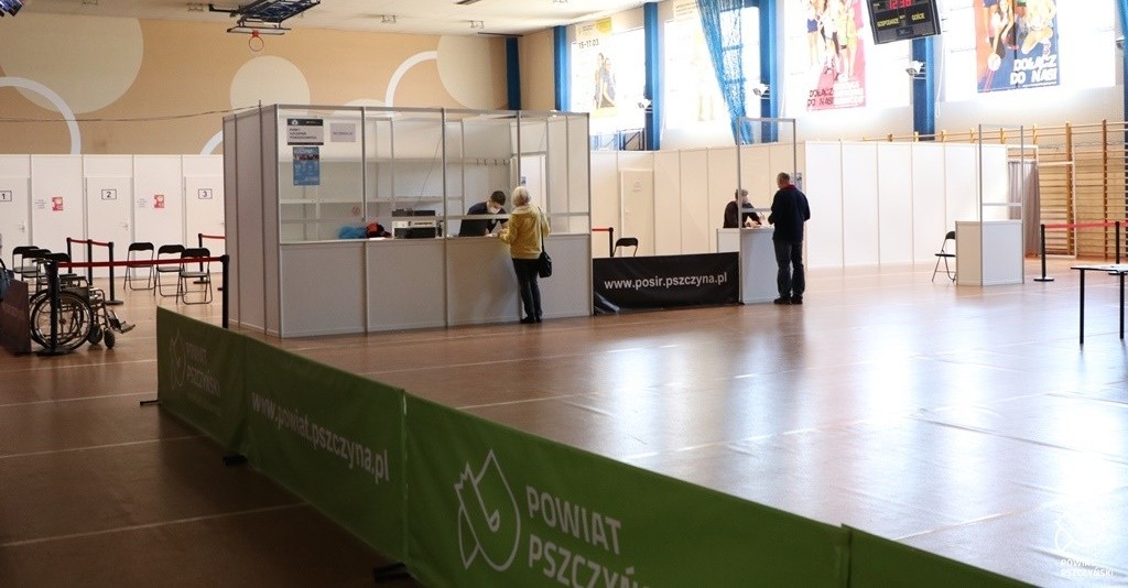 Wnętrze hali sportowej w Pszczynie, przeorganizowanej na punkt szczepień powszechnych. Na pierwszym planie zielone banery z logiem powiatu, na środku punkt informacyjny i stanowiska do szczepień.