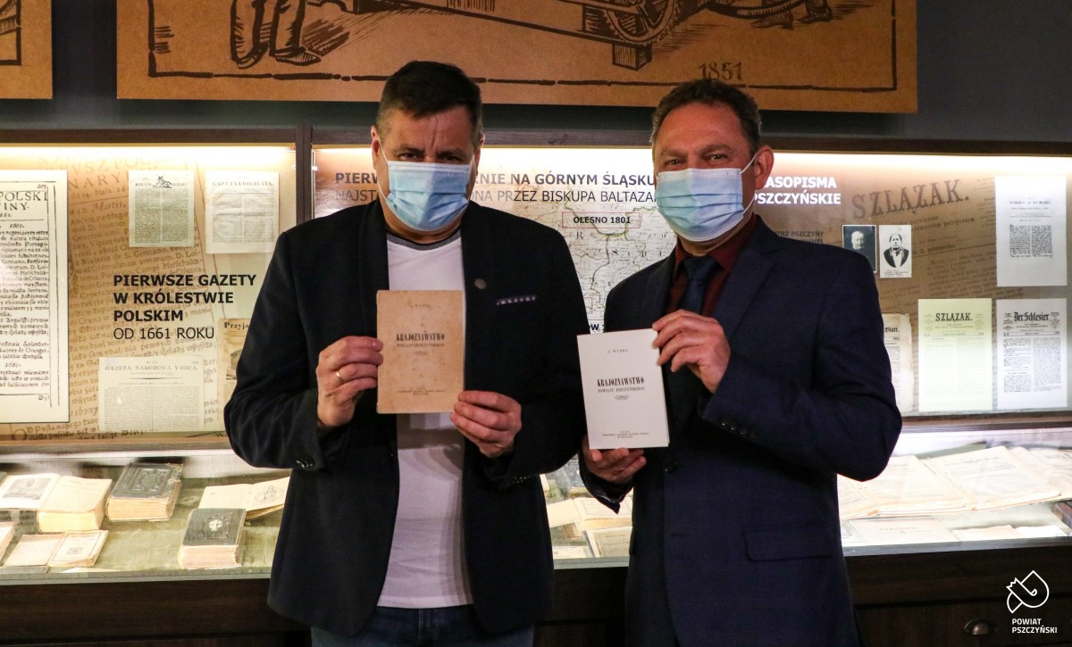 Wicestarosta Damian Cieszewski z prezesem TMZP na tle podświetlonej gabloty w Muzeum Prasy Śląskiej. W dłoniach trzymają reprint broszury.