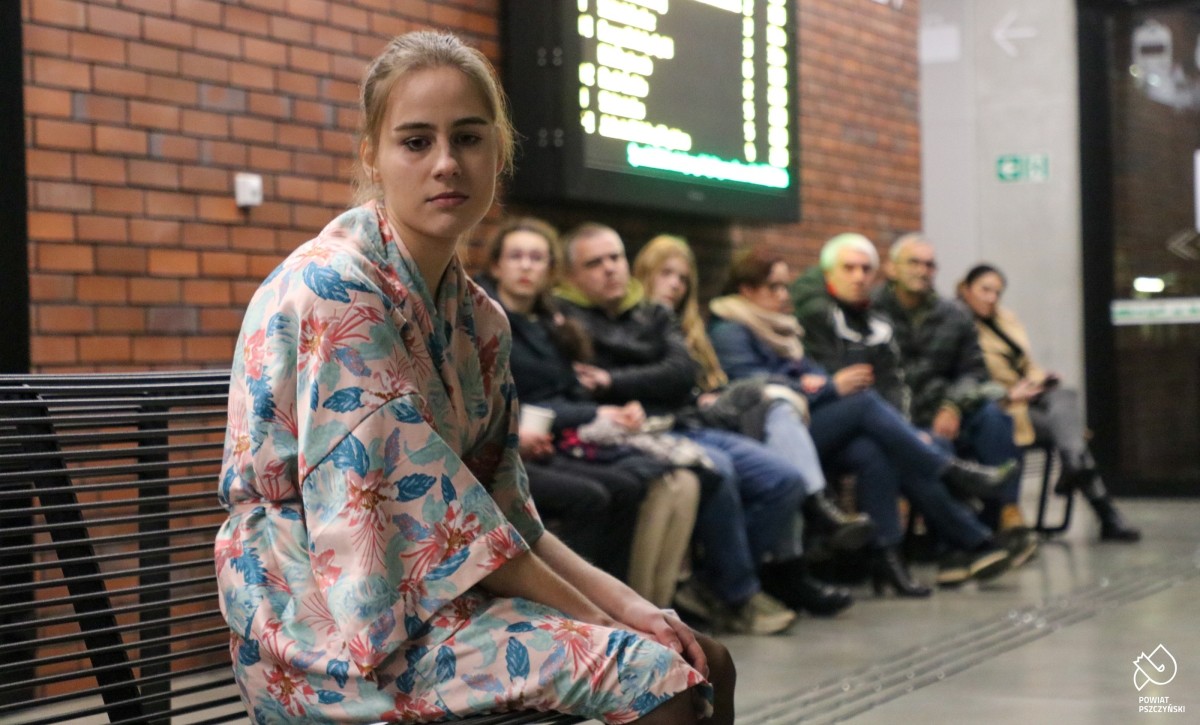 Młoda aktorka siedzi na ławce w centrum przesiadkowym. W tle widzowie.