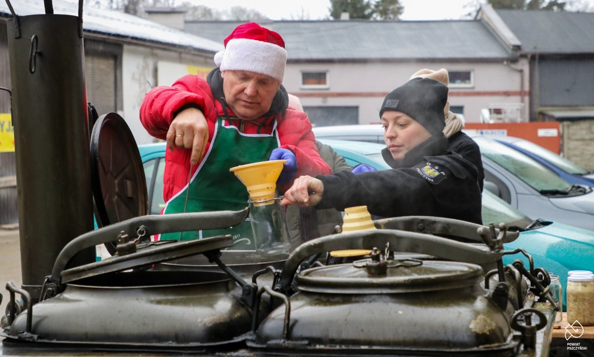 Starosta w czapce świętego Mikołaja ze strażakami nalewają zupę z kotła do słoika.