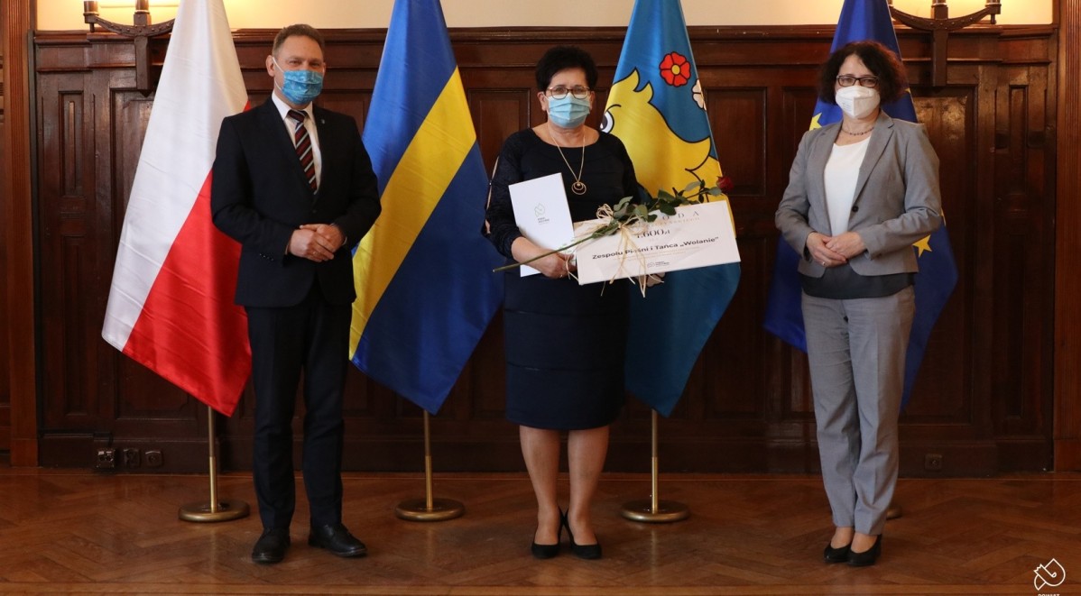 W pomieszczeniu w dużej sali na tle czterech flag stoja trzy osoby - dwie kobiety oraz męzczyzna. Trwa wręczenie nagród w dziedzinie kultury.