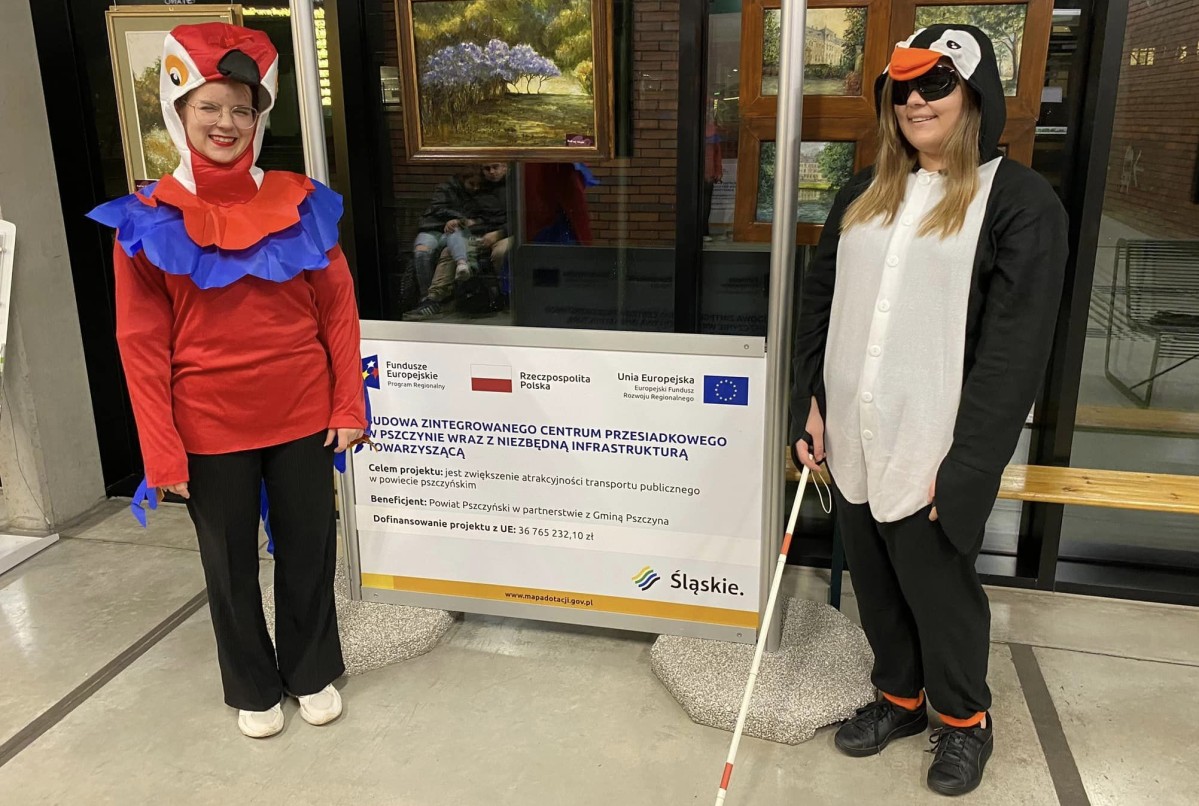 Aktorki przebrane w strój pingwina z laską oraz papugi w budynku centrum przesiadkowego.