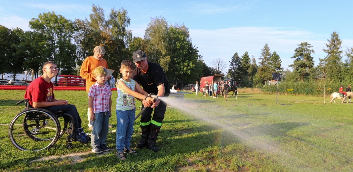 Uczestnicy pikniku z pomoca strażaka gaszą leją wodę z węża strażakiego.