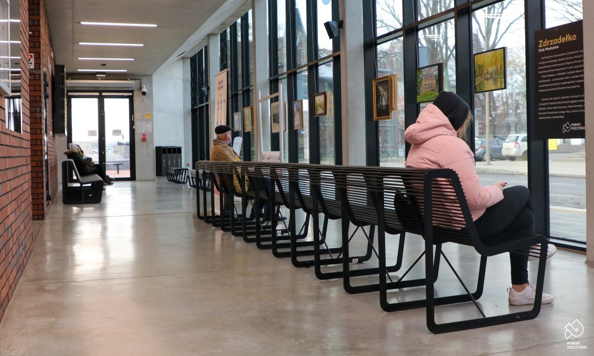 Wnętrze centrum przesiadkowego. Przed przeszkloną ścianą wiszą obrazy. Na metalowych ławkach siedzi kilka osób.