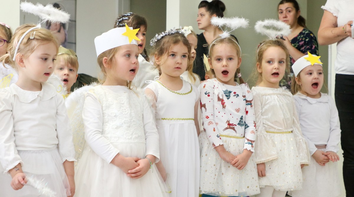 Dzieci przedszkolne ubrane w stroje aniołków stoją w pomieszczeniu i śpiewają