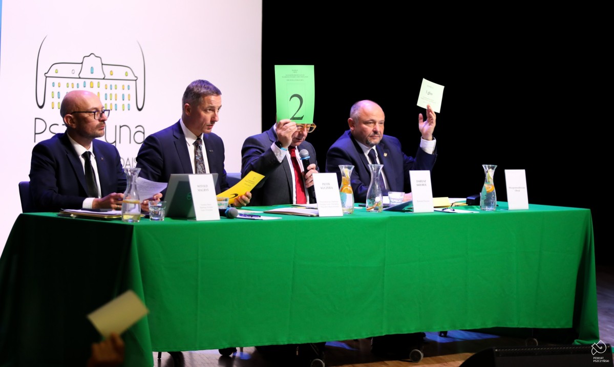 Cztery osoby przy stole z zielonym obrusem, podnoszą do góry kartki w trakcie głosowania.
