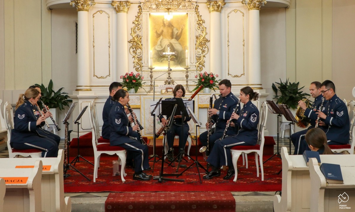 Połączeni muzycy kwartety i kwintetu grają na instrumentach wewnątrz kościoła. Siedzą na krzesłach.