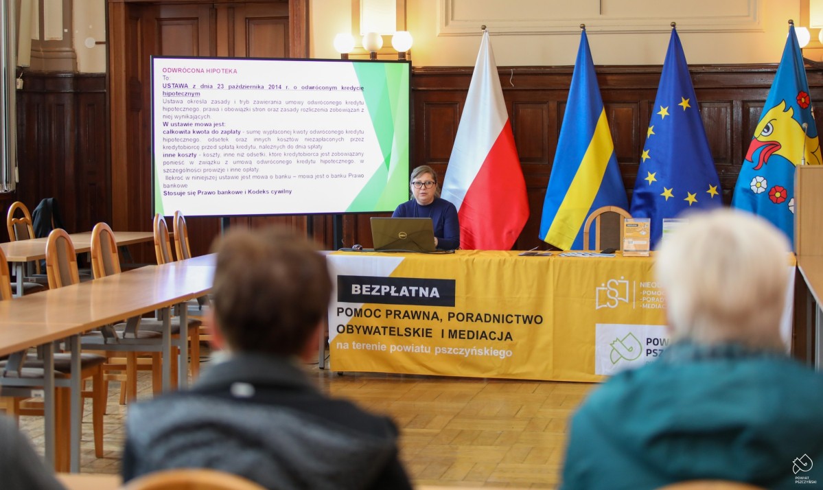 Prawniczka siedząca za stołem, za jej plecami flagi i ekran z prezentacją. Przy stołach uczestnicy.