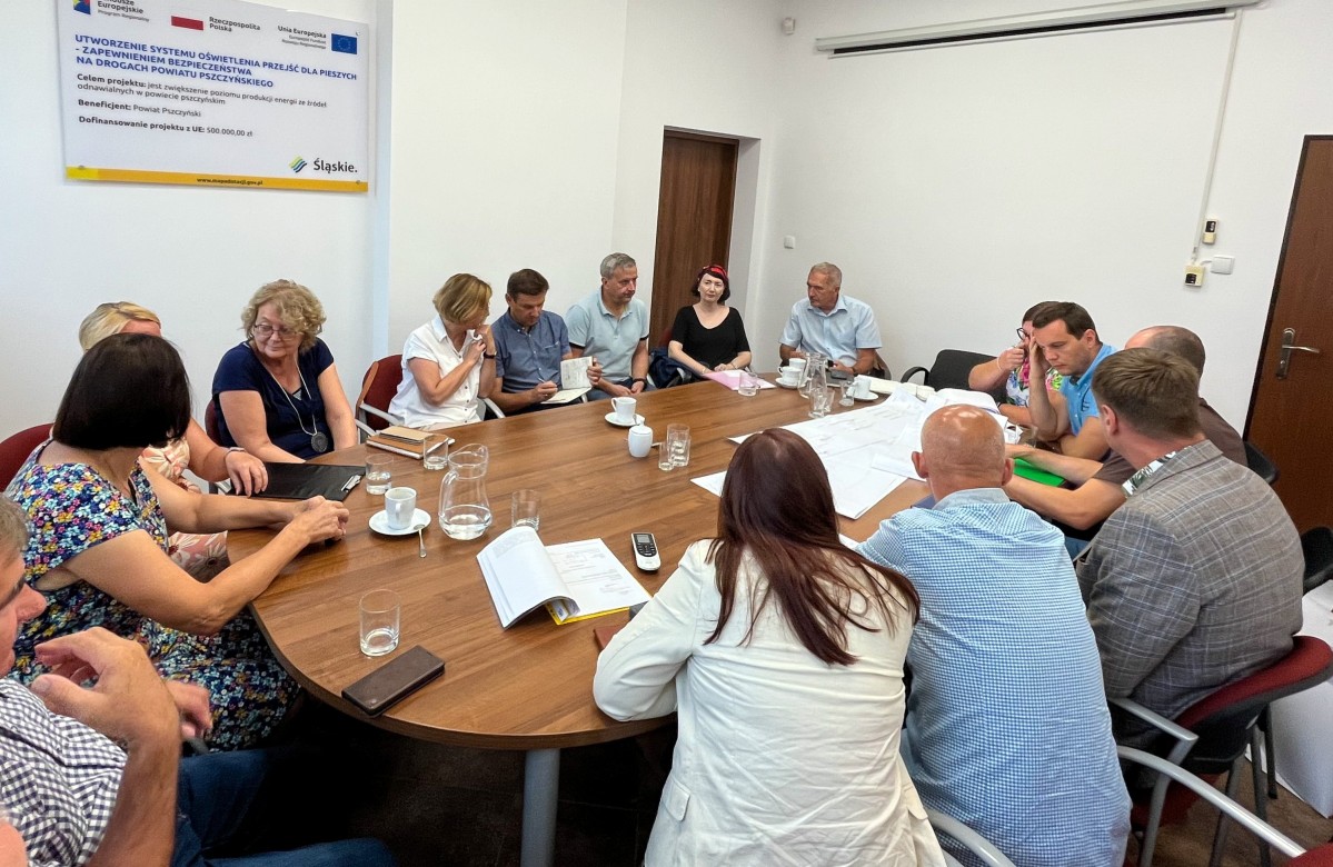 Uczestnicy spotkania siedzą przy stole, na którym położone są mapki i projekty.