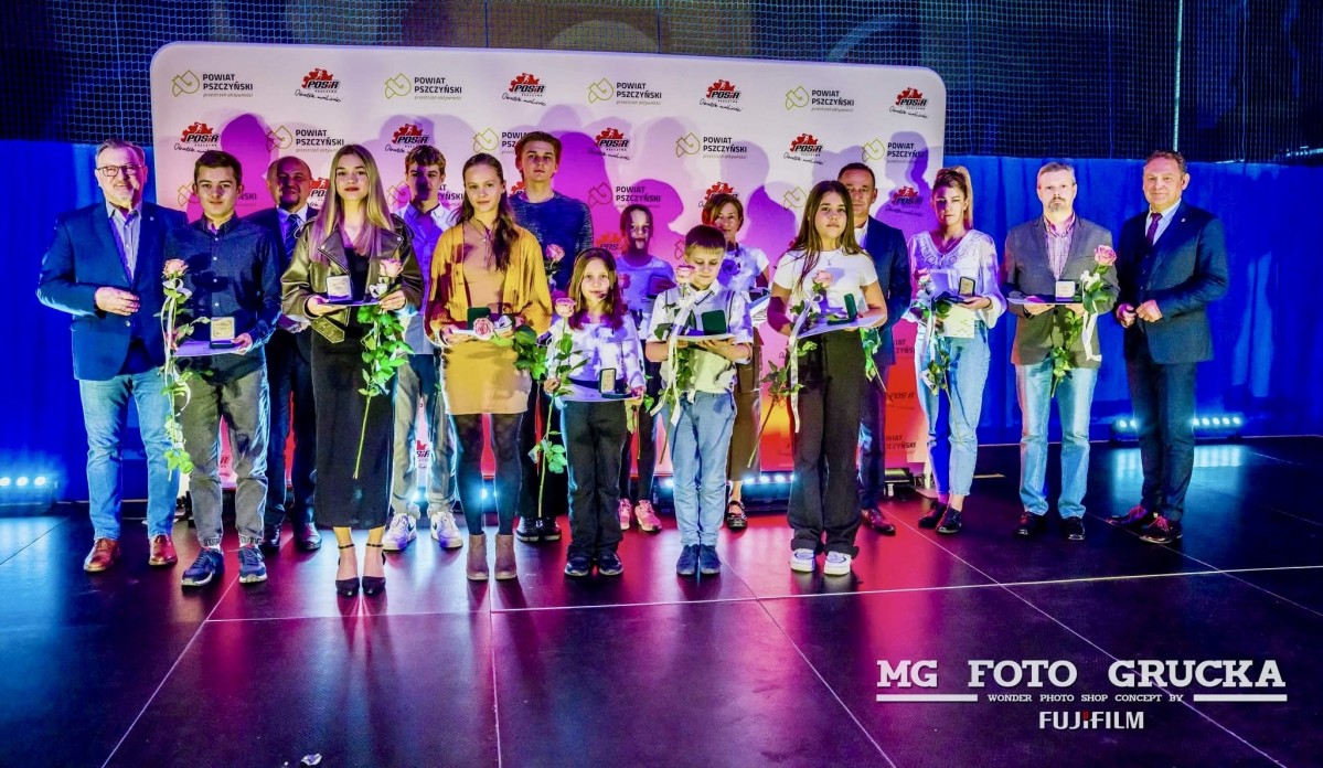 Sportowcy stoją na scenie, trzymają róże, listy gratulacyjne i nagrody.