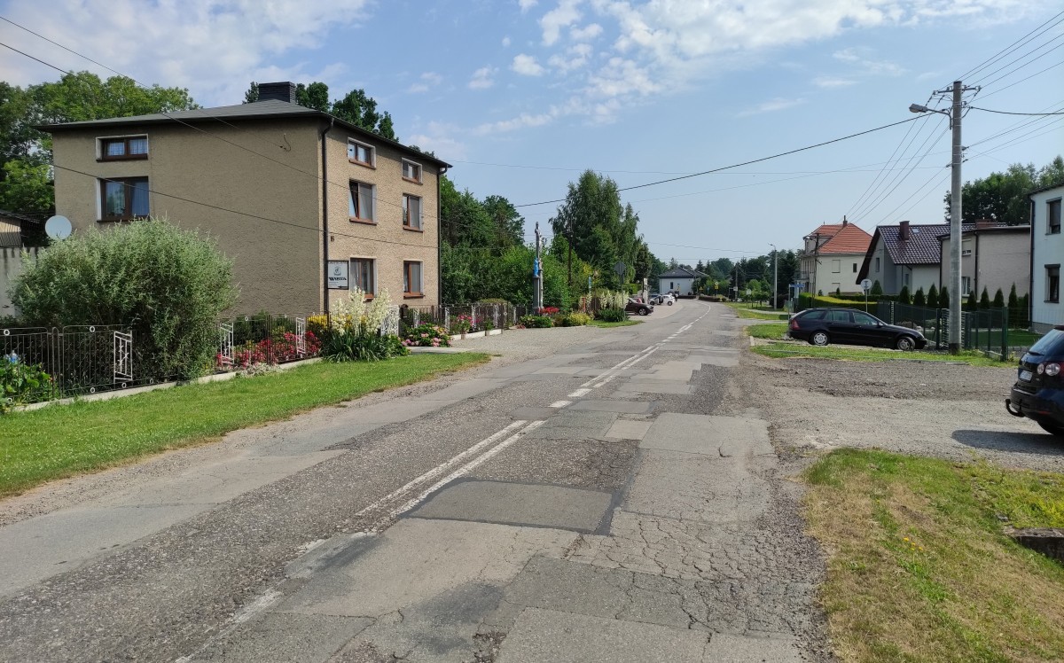 Popękana jezdnia. Wzdłuż niej zabudowa. W oddali parking kościelny. Na poboczach stoją samochody.