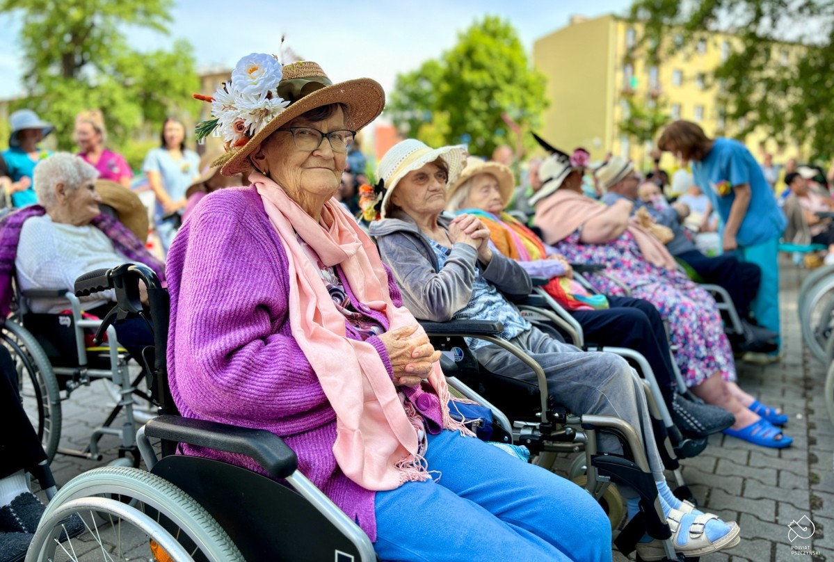 Seniorzy na wózkach siedzący przed budynkiem szpitala przy zakątku terapetycznym podczas koncertu.