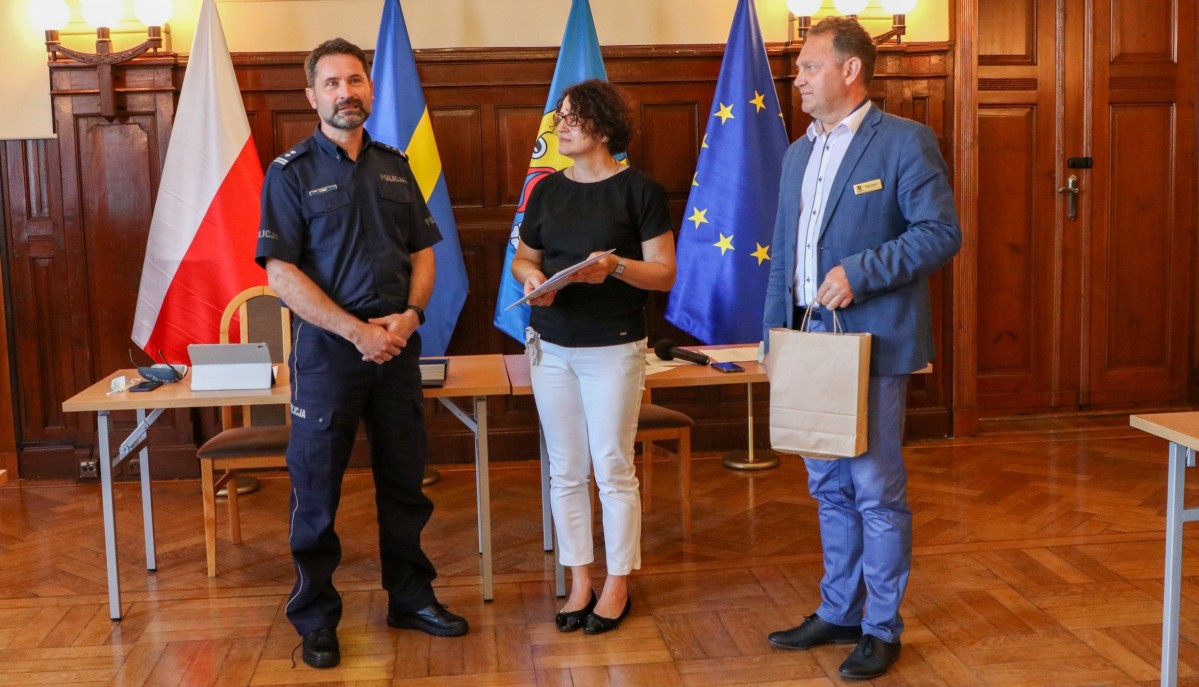 W sali sesyjnej na tle flag stoją trzy osoby - zastępca komendanta policji, starosta pszczyński oraz wicestarosta.